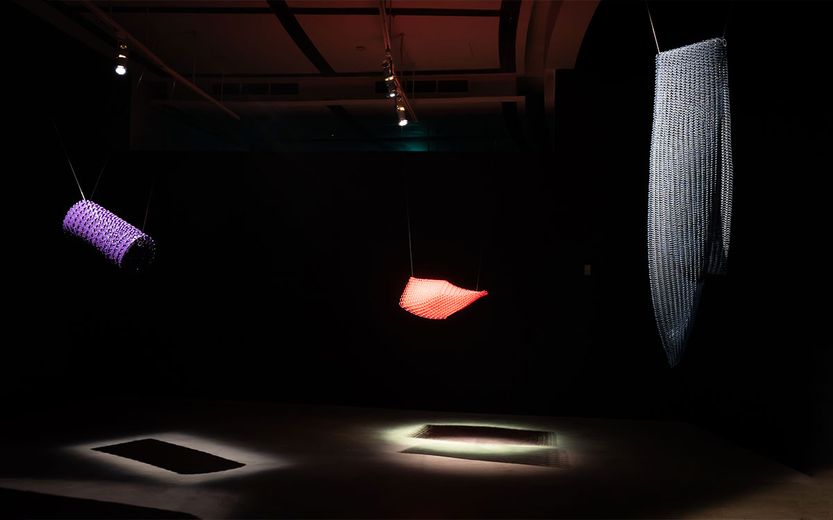 Dimensions, 2023, Grace Tan. Three crystal-like fabrics of different shapes in purple, red and grey are lit suspended against a dark background.