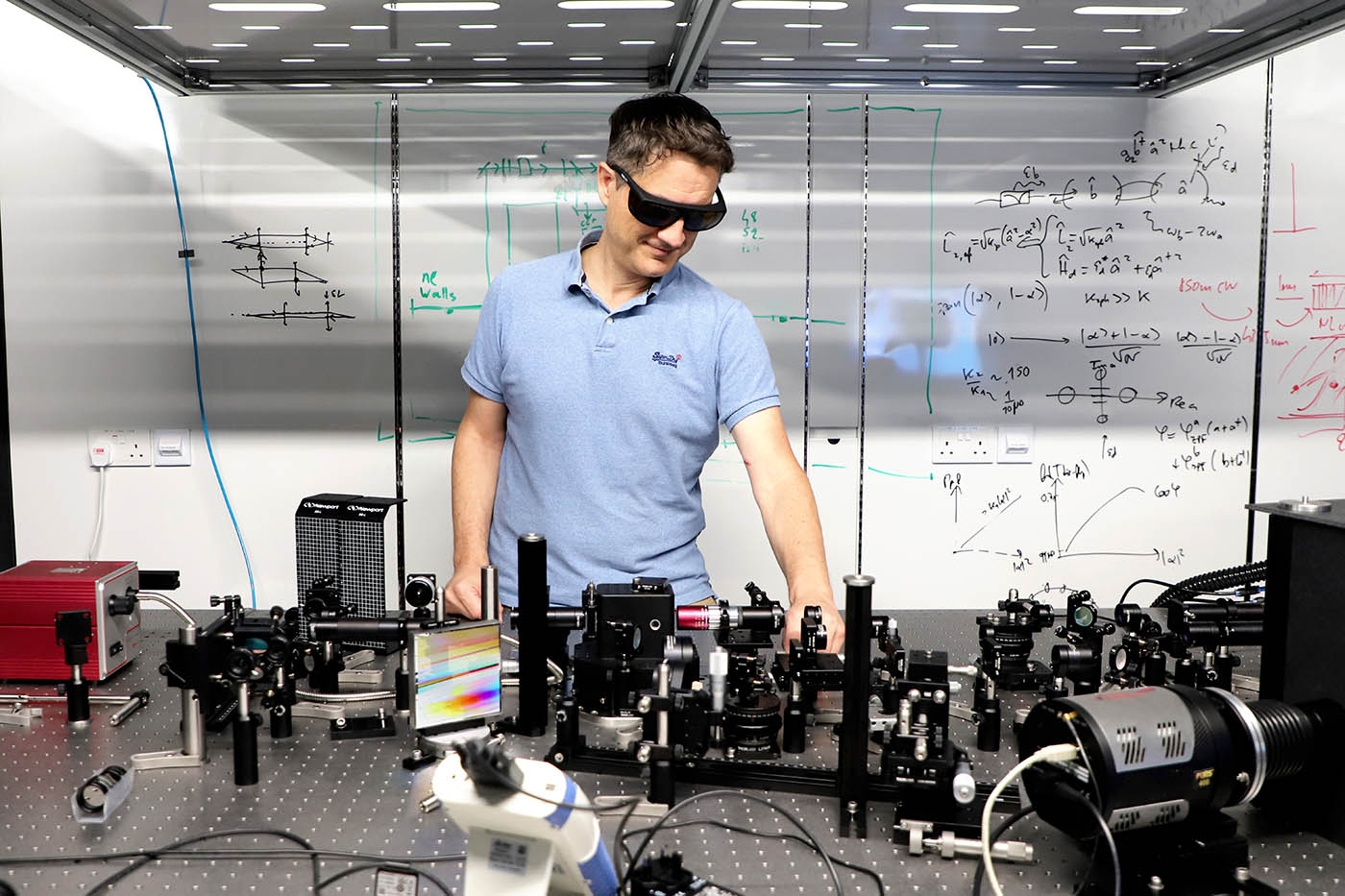 Researcher Maxime Richard stands behind a metal optical bench with components arrayed across the top. He is dressed in a pale blue polo shirt and wears laser safety glasses.