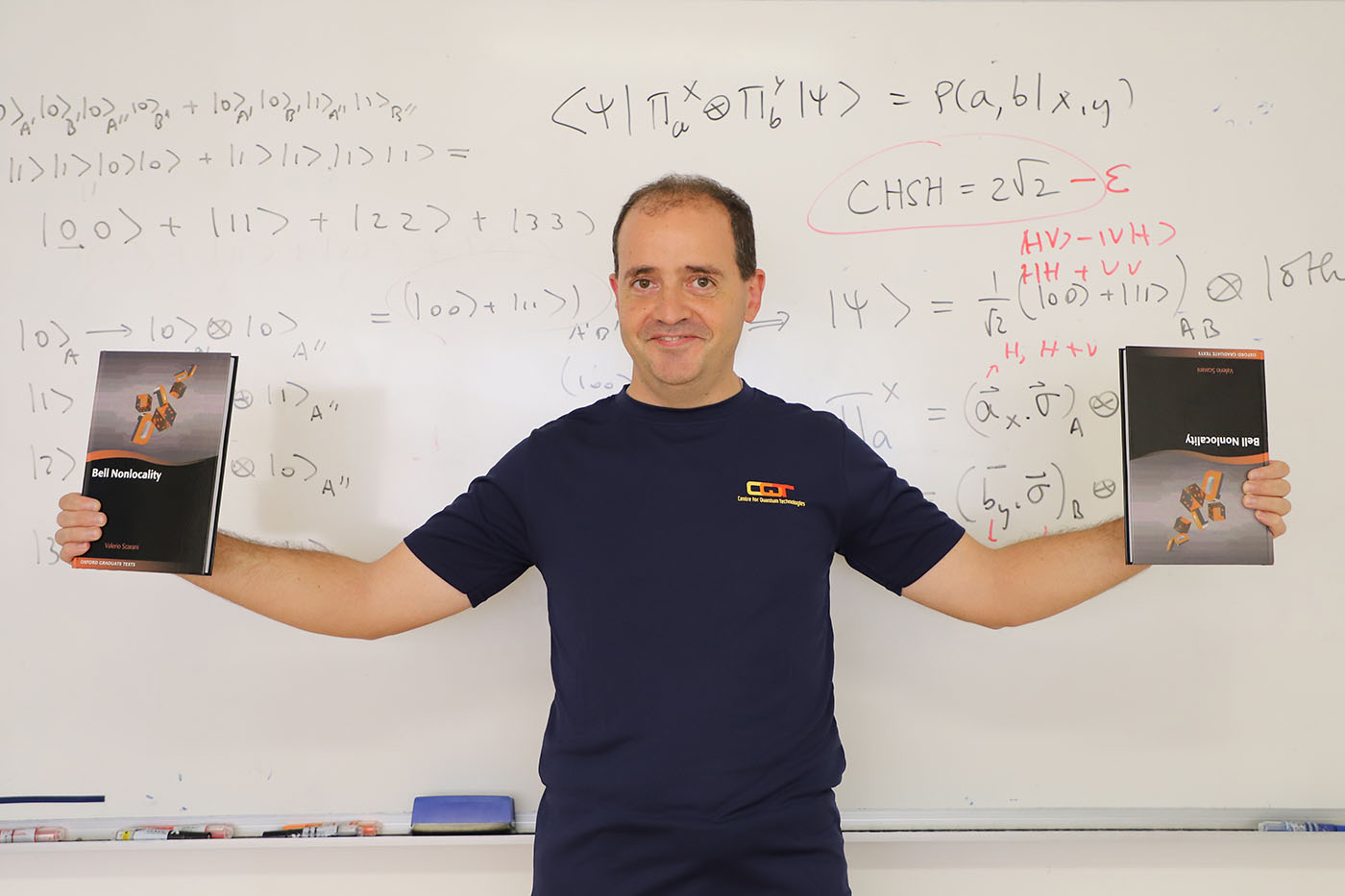 Valerio Scarani with two copies of his book in front of a whiteboard with quantum physics equations